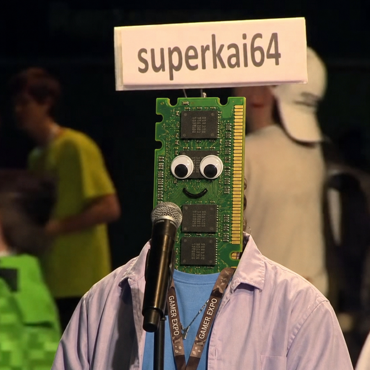 A person at a gaming expo with a RAM stick attached to their face like a mask, googly eyes and a smile drawn on it, name tag reading 'superkai64'